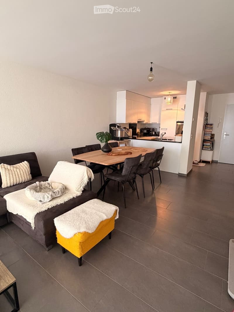 Modern living and dining area with a grey sofa, yellow stool, dining table with chairs, and an open kitchen area with white cabinets and appliances.