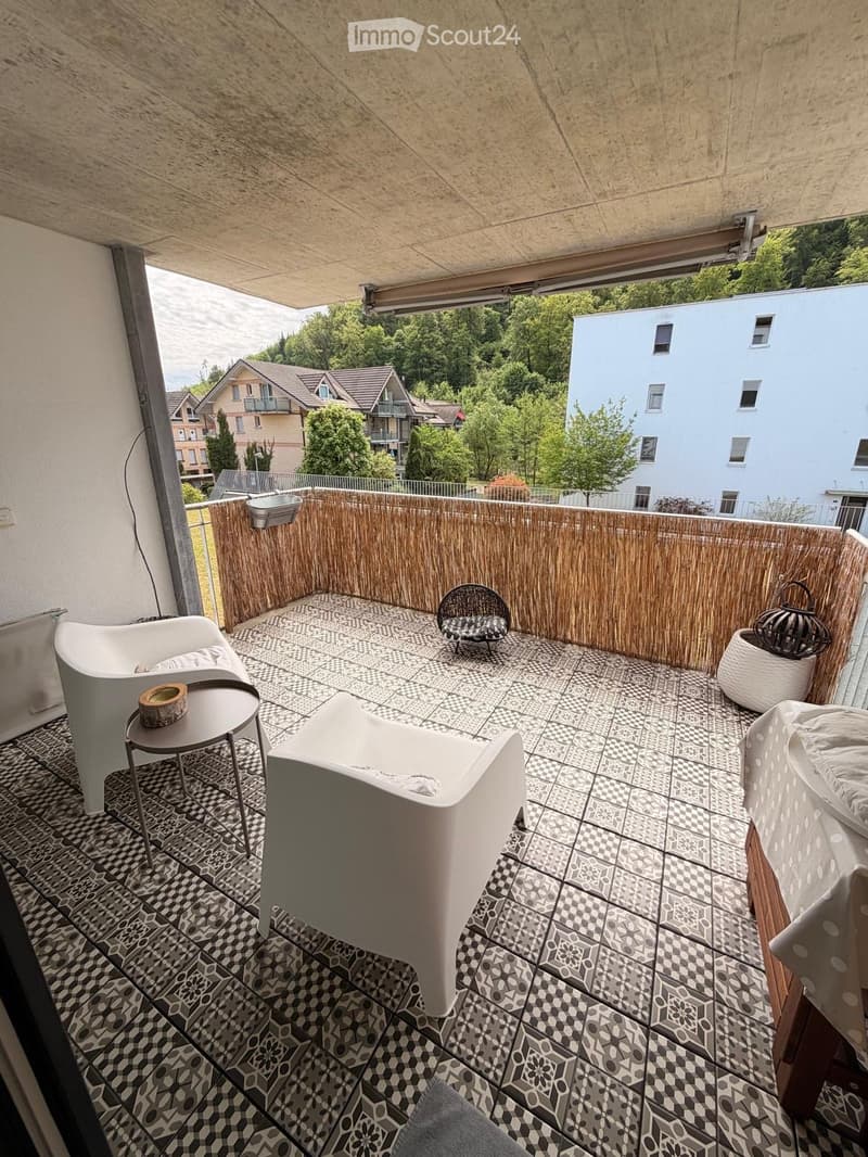 Balcony with modern furniture, including white chairs, a small table, and a wicker chair, surrounded by a bamboo fence and overlooking a green area with trees and buildings.