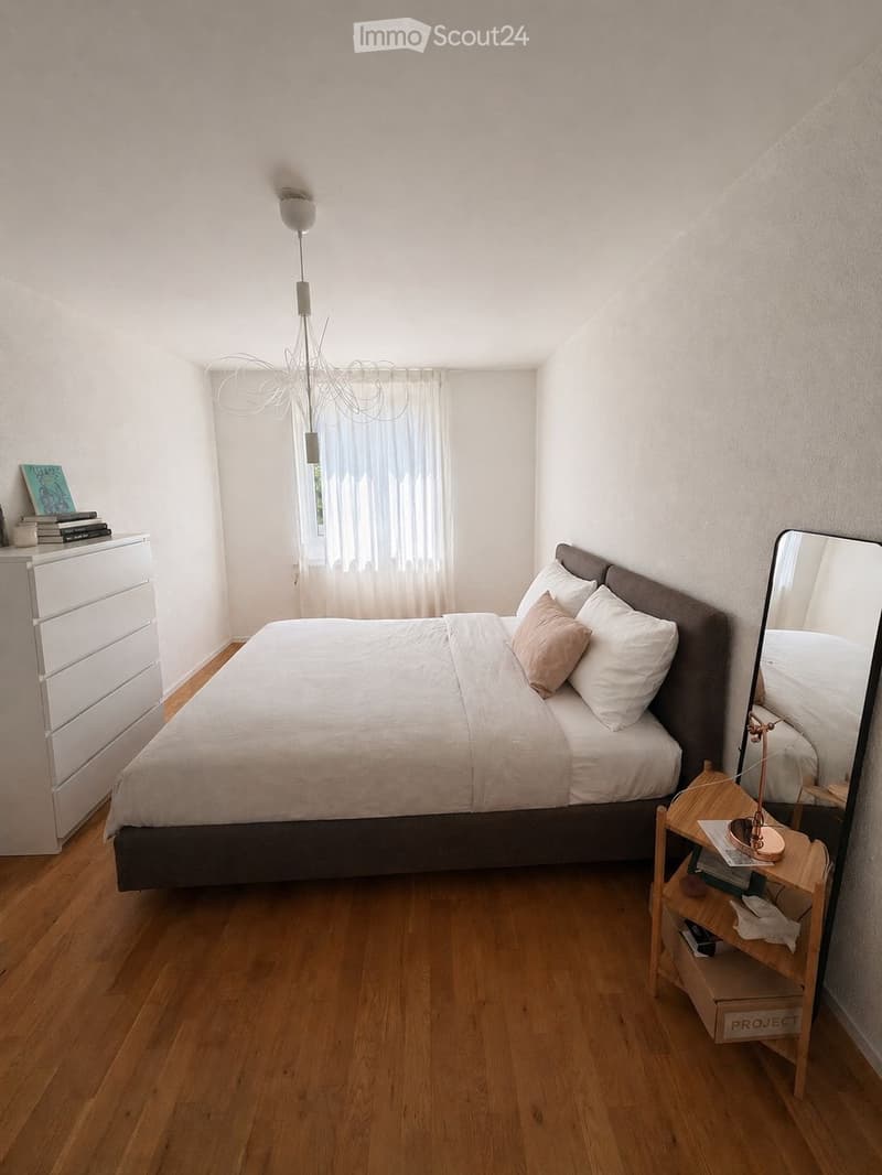A modern bedroom with a large bed, nightstand, mirror, chandelier, and a small wooden table with books and decorative items.