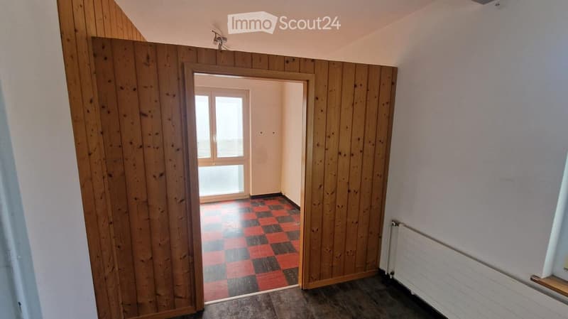 A narrow hallway with wooden panel walls, a checkered floor, and a door leading to a room with a window.