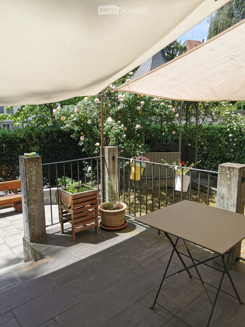 Balcony with a table, bench, potted plants, and a view of a garden with flowers and trees.