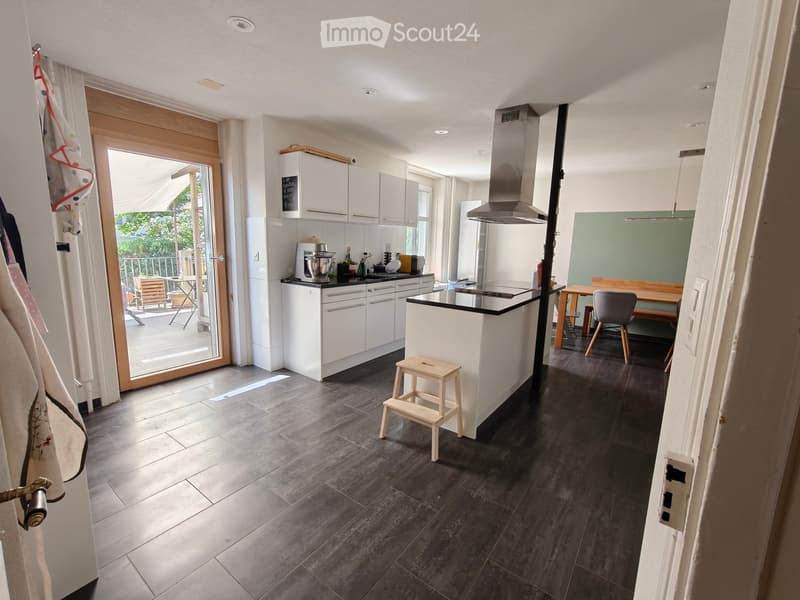 Modern kitchen with white cabinets, black countertops, and a central island. Features include a refrigerator, stove, blender, and a small wooden stool. A dining area with a table and chairs is visible, and there is a sliding glass door leading to a balcon
