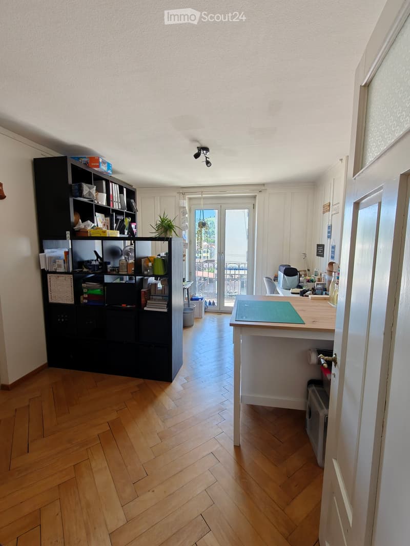 A well-lit room with wooden flooring, a large black shelving unit, a small table with a green mat, and a doorway leading to a balcony.