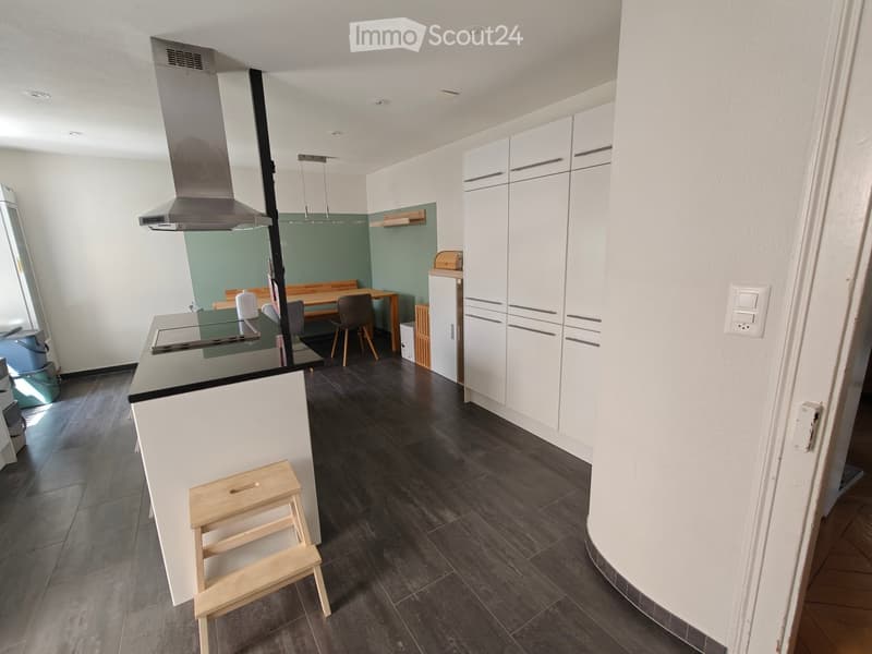 Modern kitchen with white cabinetry, dark tiled floor, kitchen island with stove, dining area with table and chairs, and a small wooden stool.