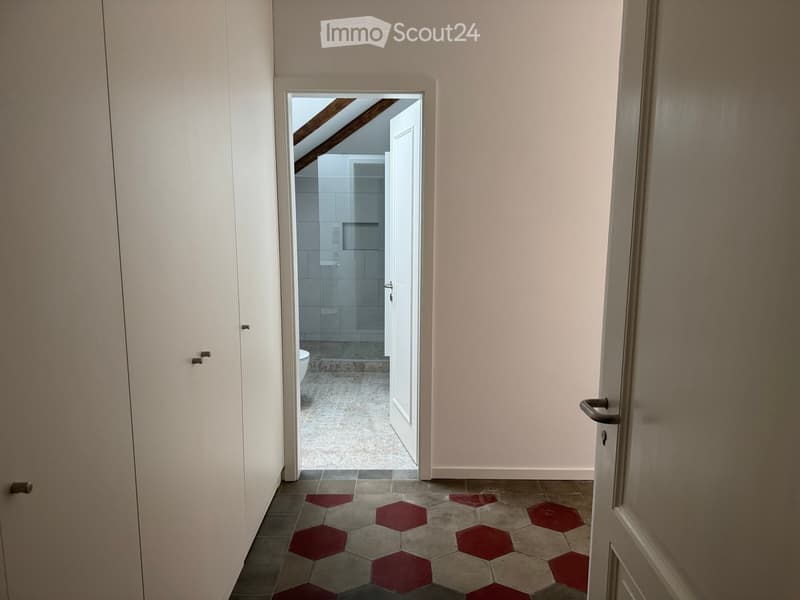 Hallway with beige walls, red hexagonal tiles, and an open door leading to a bathroom with white tiles and a toilet.