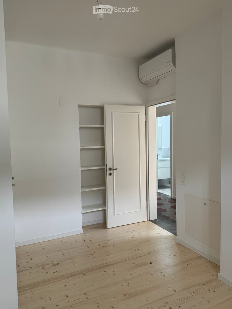 Empty room with wooden flooring, white walls, a built-in shelf unit, and an open door leading to another room.