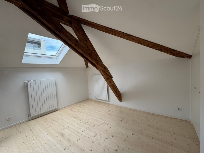 Empty attic room with wooden beams, skylight window, wooden floor, and white radiators on the walls.