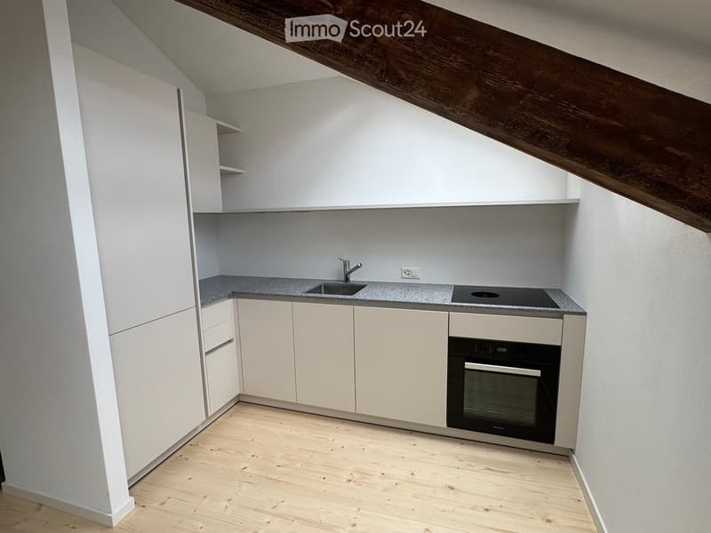 Compact kitchen with white cabinets, grey countertop, sink, faucet, and built-in oven. Wooden floor and slanted ceiling beams.