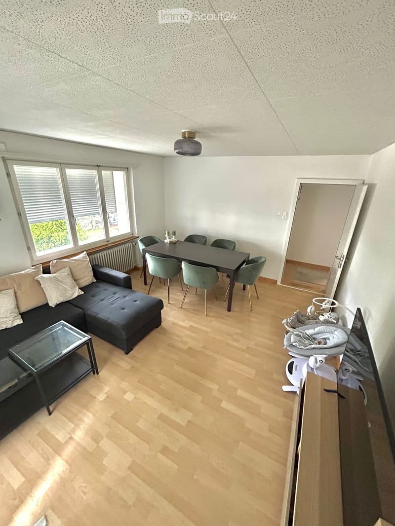 Living room with a black sectional sofa, glass coffee table, dining table with chairs, baby chair, baby seat, and a hanging lamp.