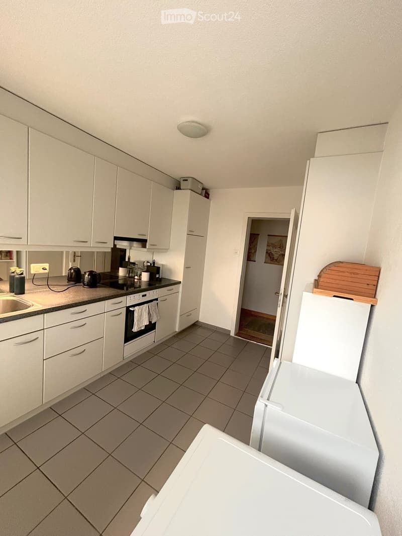 Modern kitchen with white cabinets, tiled floor, and stainless steel appliances including a sink, oven, and refrigerator. Coffee maker and oven mitts are visible on the counter.