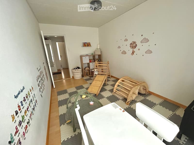 A well-lit children's room with wooden flooring, a playpen, crib, table, chair, and wall decorations including clouds and stars.