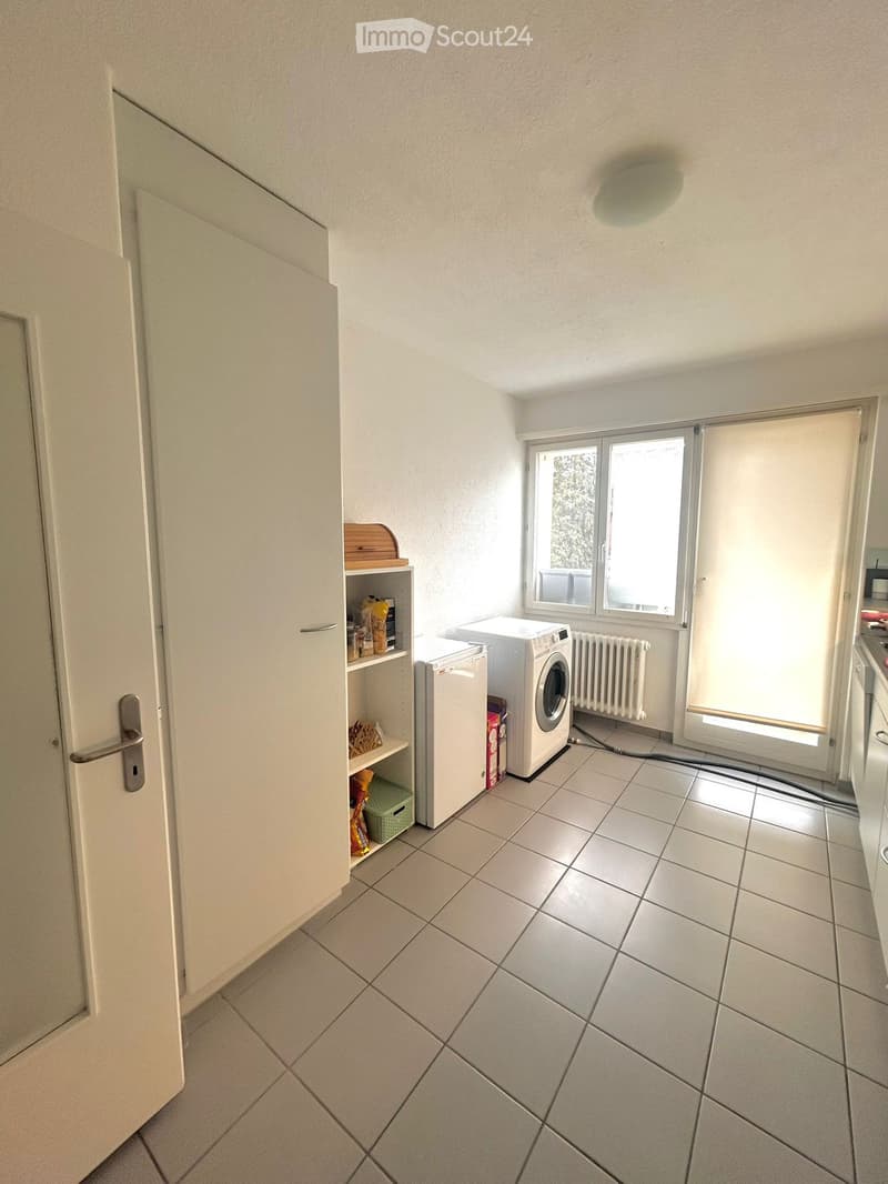 A small, tiled room with a washing machine, refrigerator, and a shelf with various items. The room has white walls, a window with blinds, and a door.