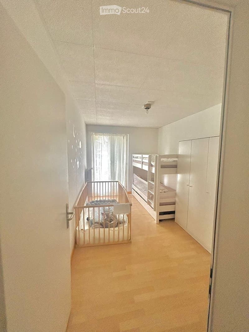 A small, well-lit room with a bunk bed and a baby crib, featuring light-colored walls and a wooden floor.