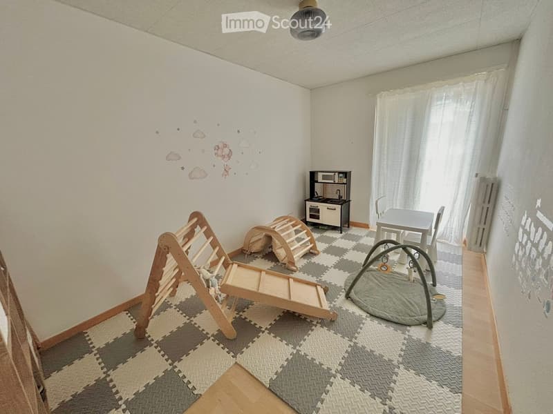 Brightly lit room with a play area featuring a wooden play structure, a play gym, a small toy kitchen, a table, and chairs. The walls are decorated with cloud and star decals.