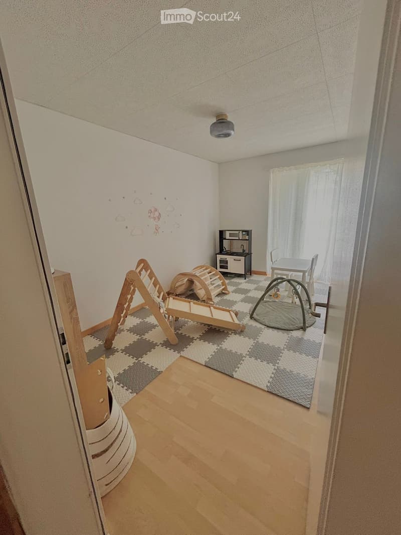 Brightly lit children's room with wooden flooring, a play area featuring a slide, tunnel, and bouncer, a small toy kitchen, and a dining table with chairs.
