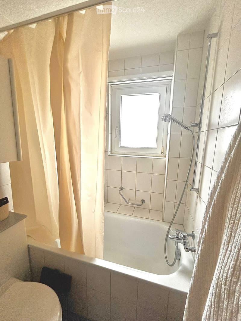 Bathtub with a handheld shower hose and a handrail, a window with a curtain, a towel hanging on the side, and a toilet visible in the corner.