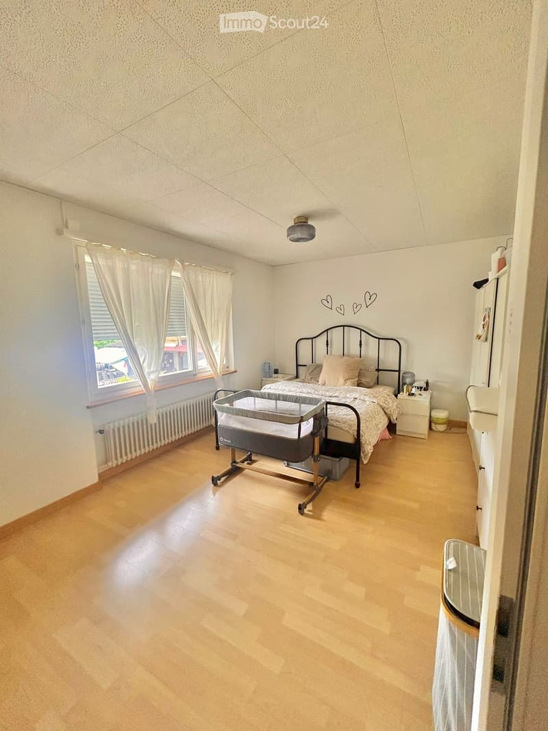 Bright bedroom with wooden flooring, large window with white curtains, bed with beige bedding, nightstand, and baby crib.