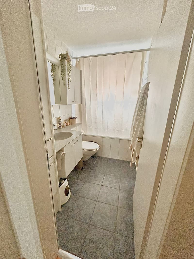 A small, well-lit bathroom with a sink, toilet, bathtub, and a window with a shower curtain. The floor is tiled, and there is a cabinet above the sink.