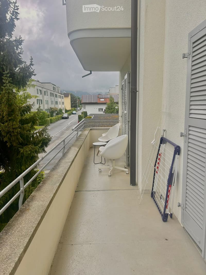 Balcony with two white chairs and a small table, overlooking a street with buildings and trees.