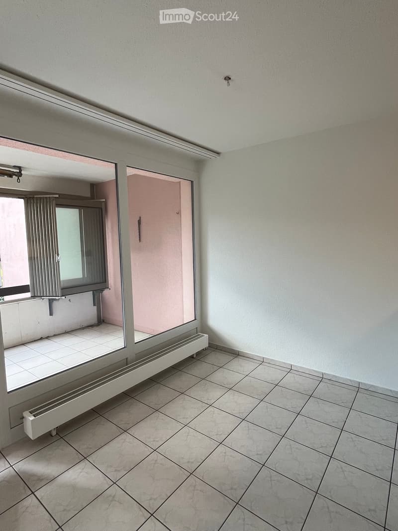 Empty room with tiled floor, white walls, and a large window with radiator below. Adjacent room visible through doorway.