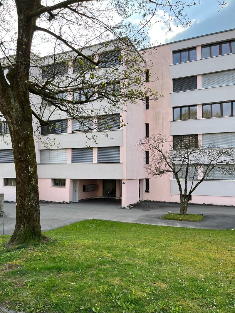 Modern two-story building with pink and white facade, large windows, and a spacious driveway. The building is surrounded by a well-maintained lawn and trees.
