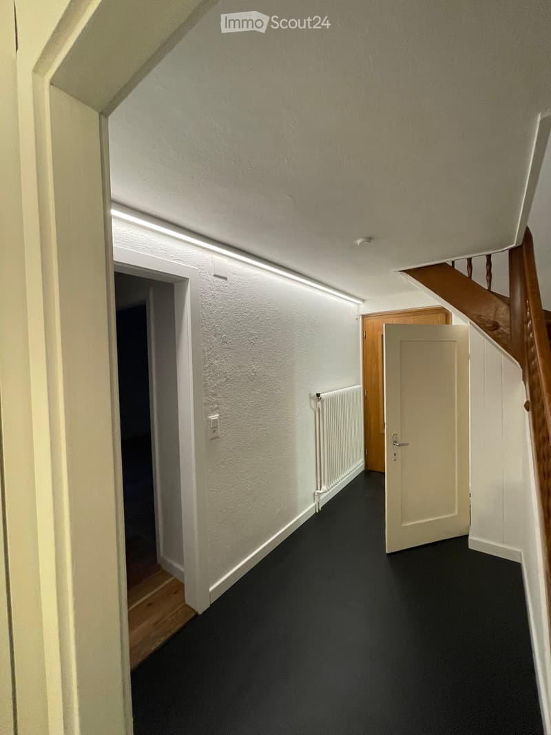 A narrow hallway with white walls, black carpeted floor, and a wooden staircase leading upwards. There are doors on both sides and a radiator on one wall.