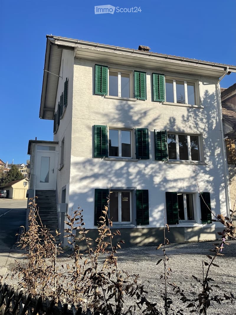 Two-story white house with green shutters, multiple windows, and a small entrance door. The house is surrounded by gravel and some dry plants.