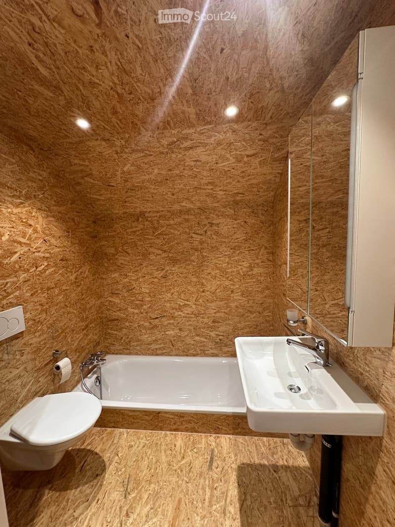 Modern bathroom with wooden walls and floor, featuring a toilet, bathtub, sink, and mirror.
