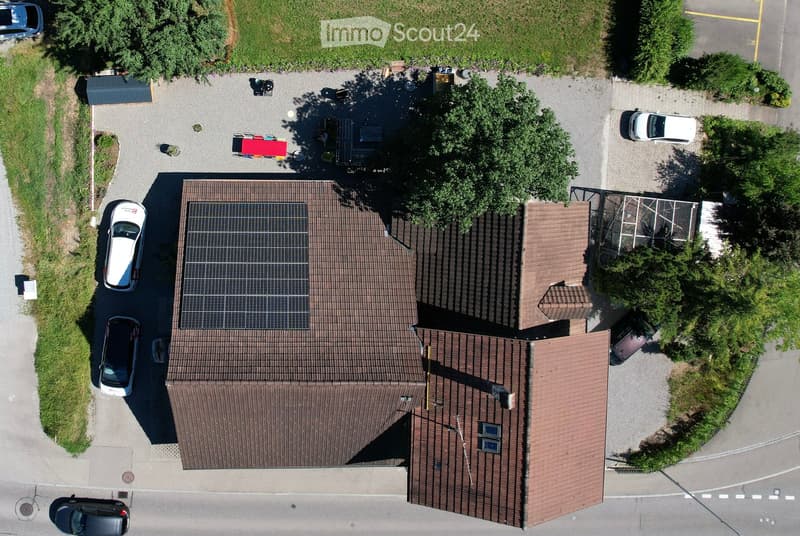 Aerial view of a two-story house with a brown tiled roof, solar panels on the roof, and a parking area with several cars.