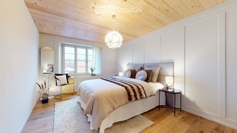 Bright bedroom with wooden floors, white walls, and a large window. Features a neatly made bed with pillows and a blanket, a nightstand with a lamp, a chair with a cushion, and a mirror on the wall.