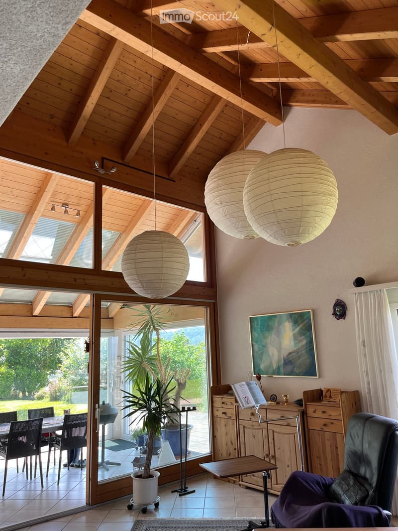 Bright living room with wooden beams, large windows, hanging paper lanterns, a leather chair, a small table, a potted plant, and a view of the garden.