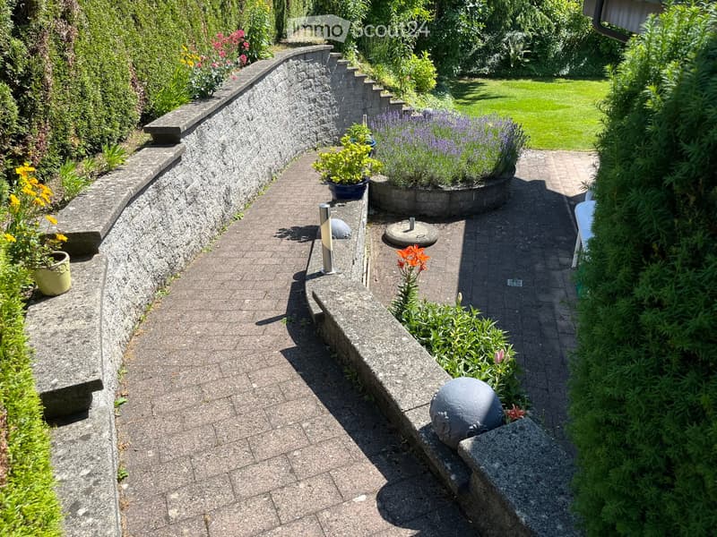 A curved garden pathway lined with stone walls, potted plants, and decorative spheres, leading to a grassy lawn.