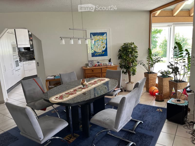 Dining area with a round table, six chairs, and a kitchen area with white cabinets, microwave, and oven. There are potted plants, a decorative ball, and a picture frame on a side table.