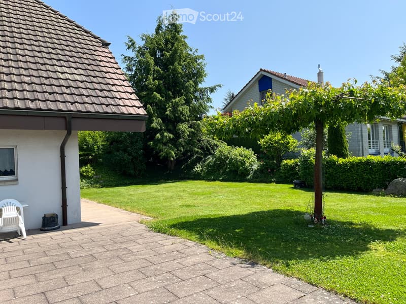 Well-maintained garden with a paved pathway, a white plastic chair, a tree, and surrounding greenery.