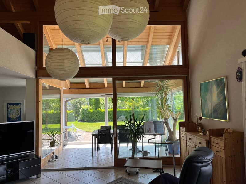 Bright living room with large windows and sliding glass doors leading to a patio. Features a sofa, coffee table, chairs, potted plants, a television, and decorative items.