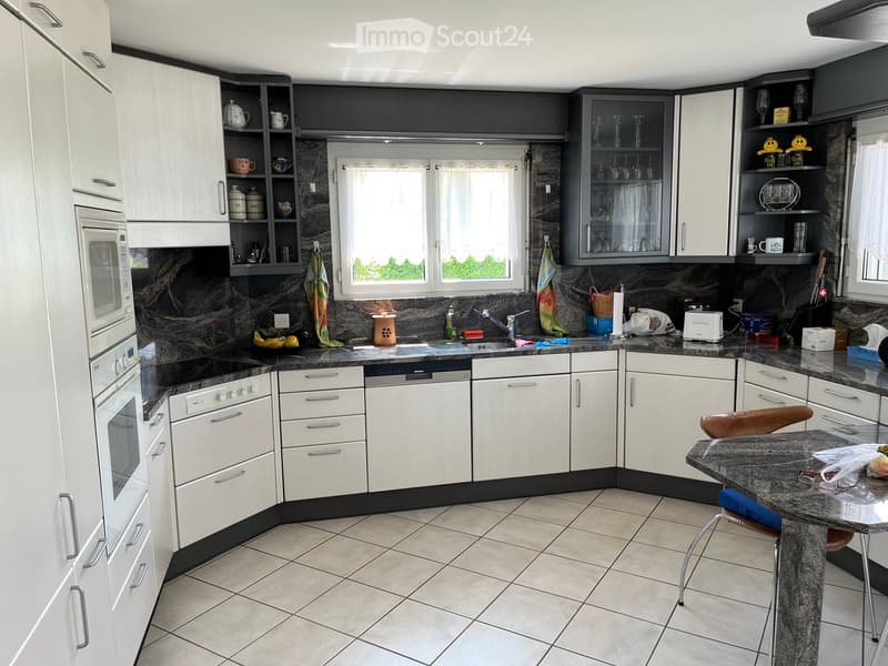 Modern kitchen with white cabinets, granite countertops, and stainless steel appliances including a microwave, oven, dishwasher, and refrigerator. Features a central island with a stool, a sink with a faucet, and various kitchen items on the counters.