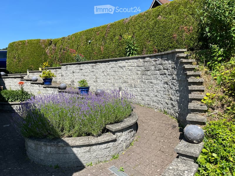 Well-maintained garden with a stone wall, lavender plants, potted flowers, decorative spheres, and neatly trimmed hedges.
