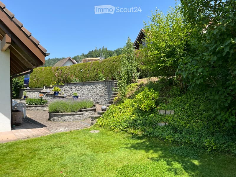 Well-maintained garden with a stone wall, steps, various plants, shrubs, and a small seating area with a white chair and table.