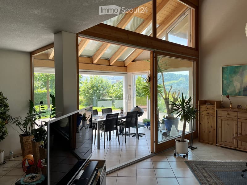 Bright living space with large windows and a view, featuring a dining area with a table and chairs, potted plants, a cabinet, and a TV screen.