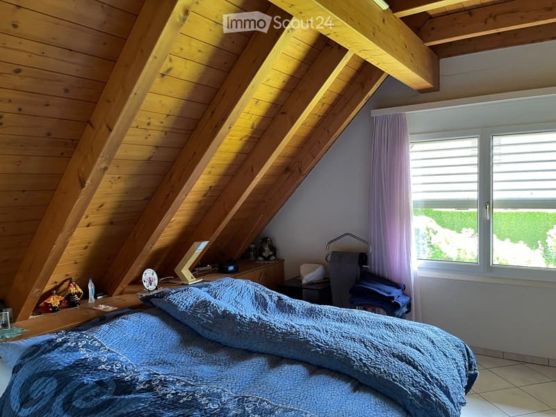Cozy attic bedroom with wooden slanted ceilings, large windows with purple curtains, a neatly made bed, nightstand with a table lamp, and decorative items on the dresser.