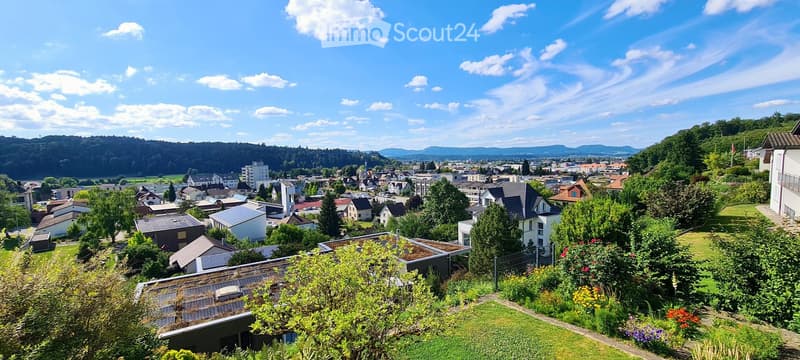 Panoramic view of a town with houses, trees, and distant mountains under a clear blue sky.