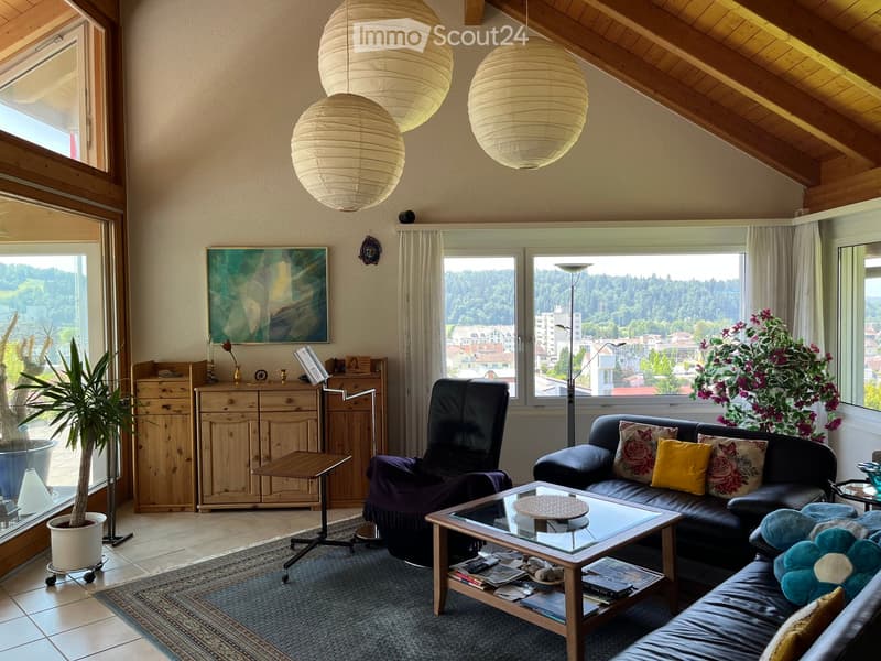 Cozy living room with exposed wooden beams, large windows offering a scenic view, a black leather sofa with colorful cushions, a glass coffee table, an armchair, a wooden cabinet, and a potted plant.