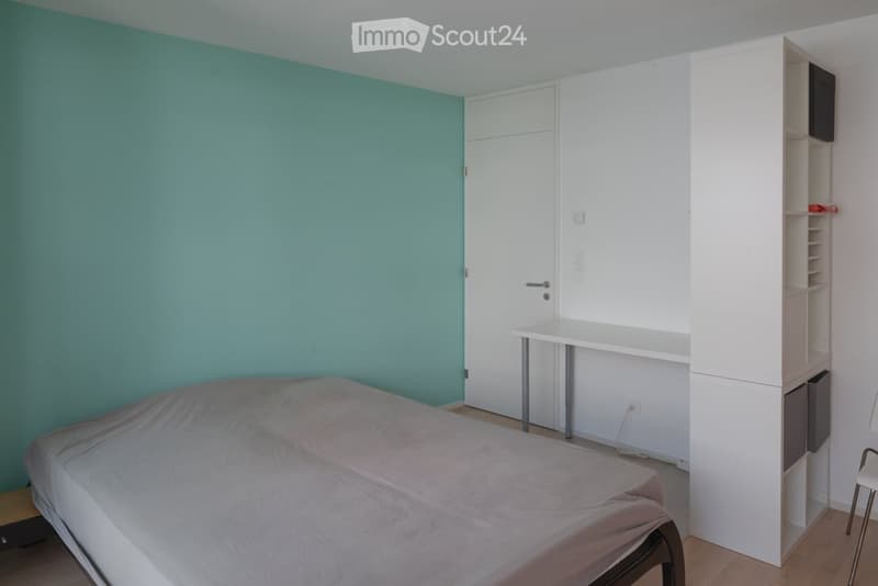 Modern bedroom with a bed, desk, chair, and built-in wardrobe. The walls are painted in a calming teal color, and the floor is wooden.