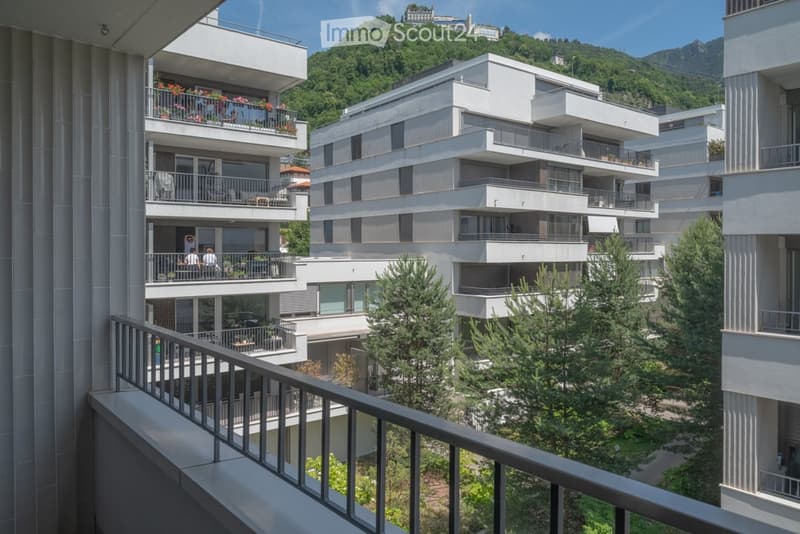 Modern multi-story residential complex with balconies, overlooking a lush green hillside and distant mountains.