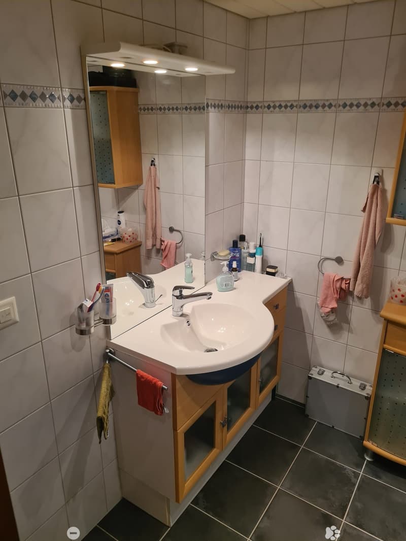 Modern bathroom with a white sink, dual faucets, a large mirror, wooden cabinets, and various toiletries on the counter. Towels are hanging on the sides.