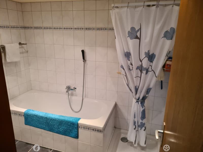 Bathroom with a white bathtub, blue towel, shower curtain with floral design, and tiled walls.