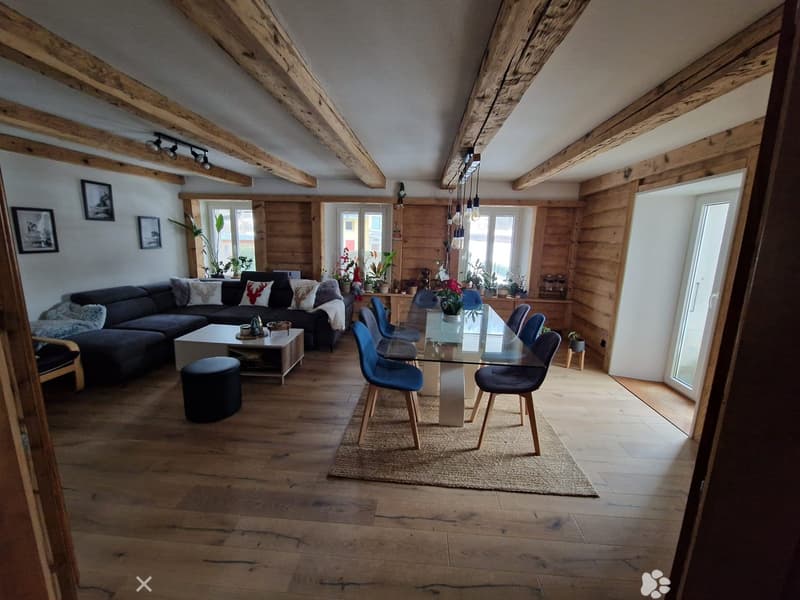 Open-plan living and dining area with wooden beams, exposed wooden floor, large windows, sofa, coffee table, dining table with chairs, and decorative items.