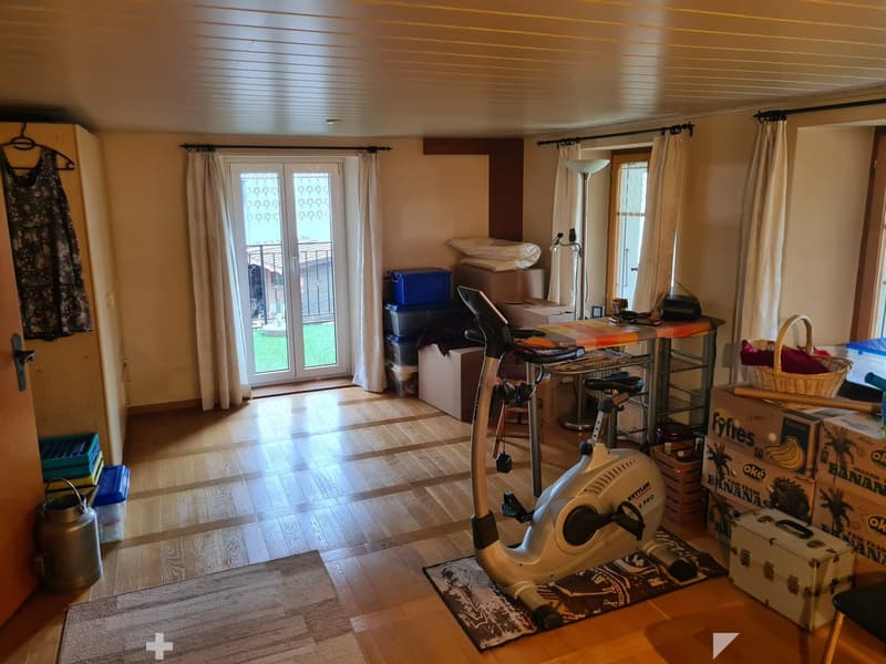A room with wooden flooring, an exercise bike, a table with a chair, a curtained sliding door leading to a balcony, and several storage boxes and baskets.