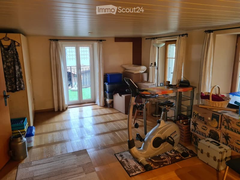 A room with wooden flooring, an exercise bike, a table with a chair, a curtained sliding door leading to a balcony, and several storage boxes and baskets.