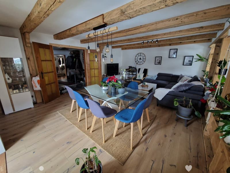 Cozy living and dining area with exposed wooden beams, hardwood flooring, a grey sectional sofa, a glass dining table with four blue chairs, and several potted plants.
